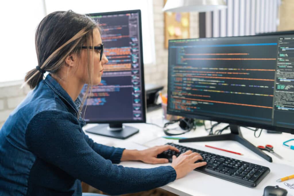 Female cybersecurity analyst working on dual monitors, analyzing code and securing networks.